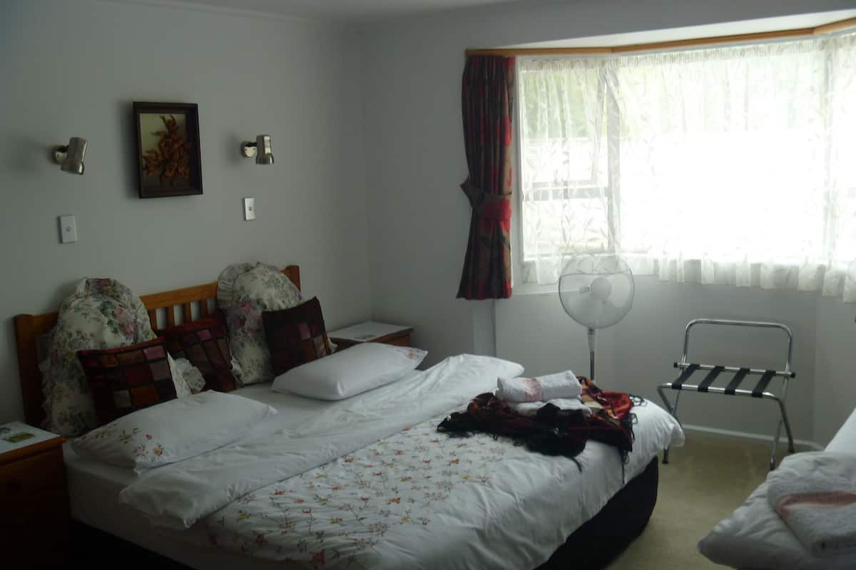 A bedroom with a bed, a bedside table, a chair, a standing fan, and a window with curtains.