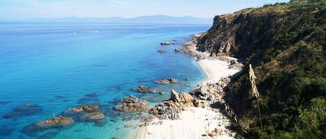 A coastal landscape with a clear blue sea, rocky shoreline, and a sandy beach.
