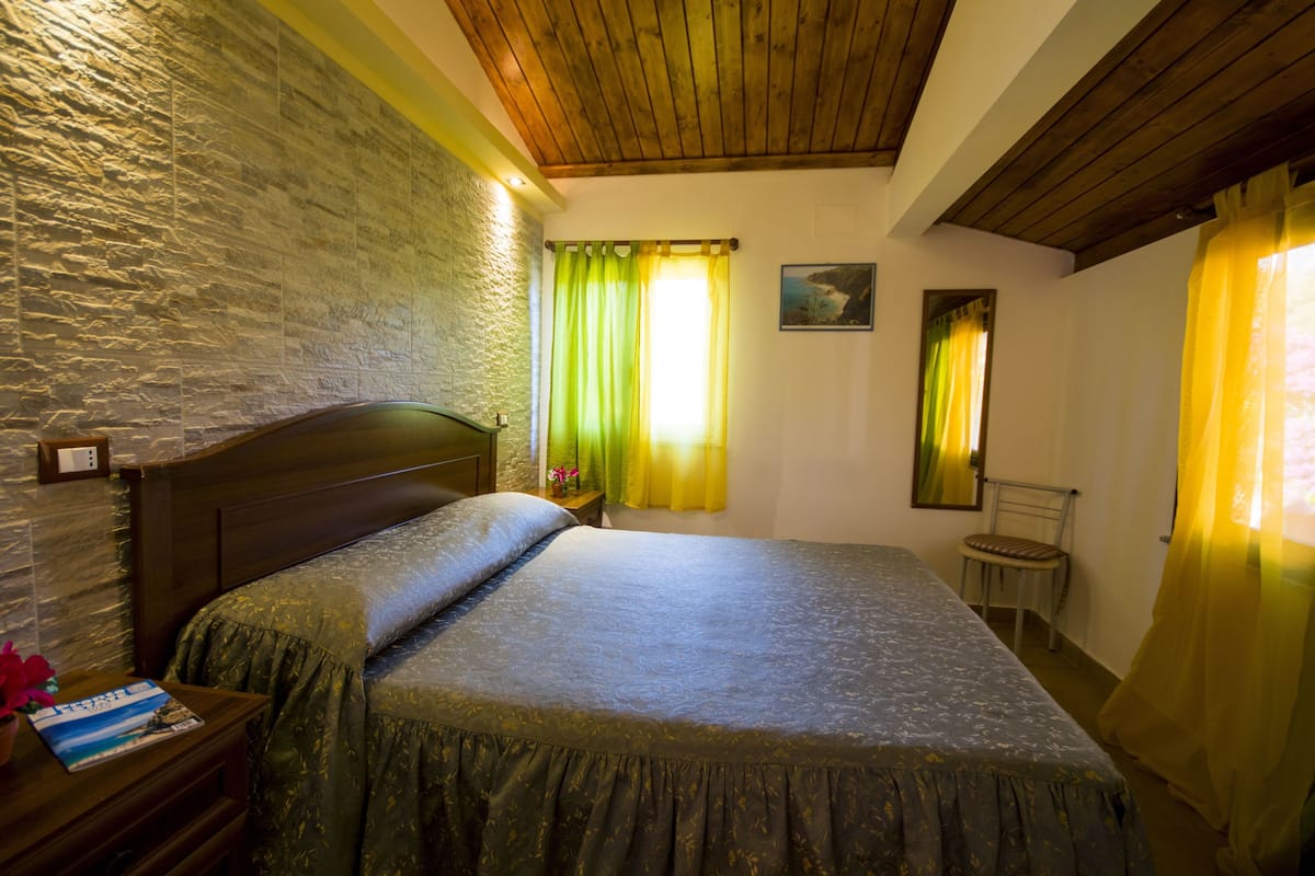 Ein Schlafzimmer mit einer Steinwand, einer Holzbalkendecke, einem Bett mit grüner Bettdecke, einem Nachttisch mit einem Buch und einem Sessel.