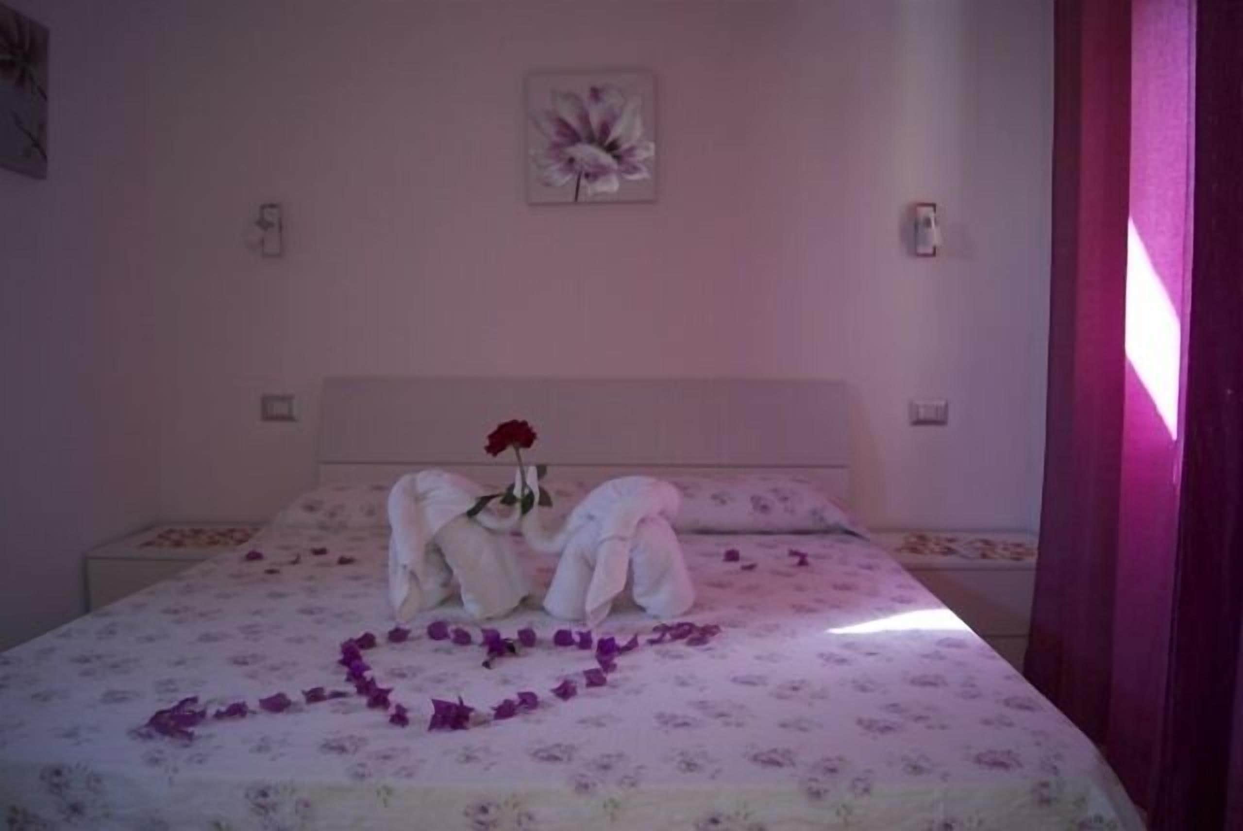 A bedroom with a floral bedspread, a heart-shaped arrangement of rose petals, and a red rose on a pillow.