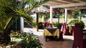 An outdoor patio with tables and chairs, surrounded by plants and a thatched roof structure. - Park Oasi Residence (Zambrone)