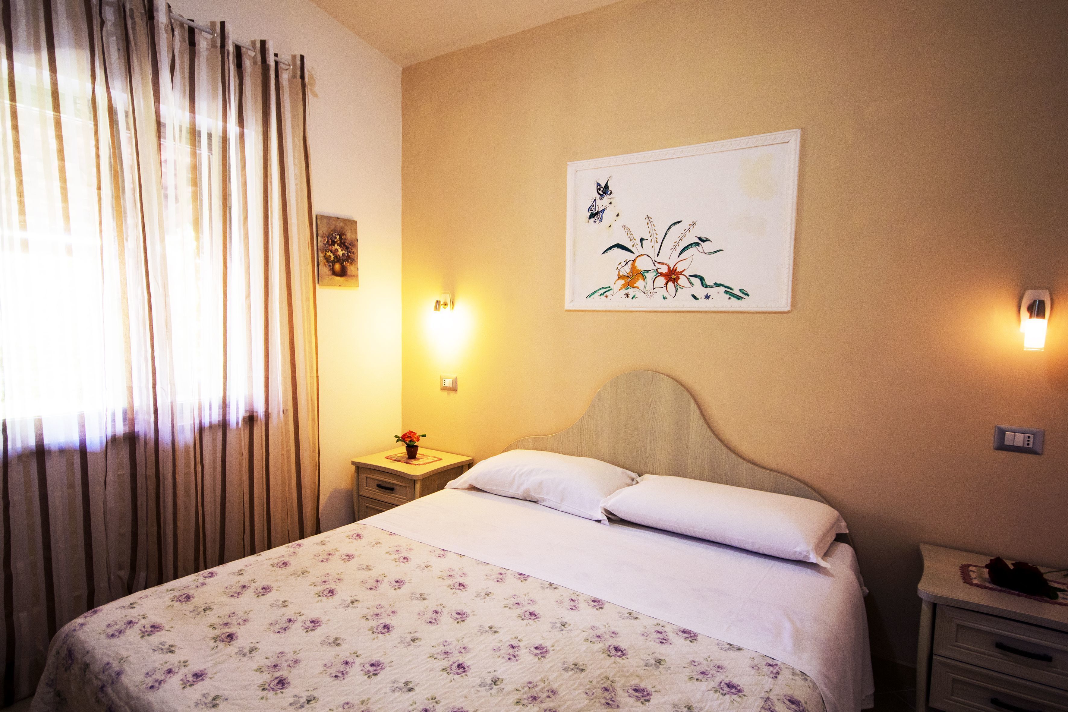 A hotel room with a bed, a nightstand with a lamp, a wall-mounted painting, and a window with curtains.