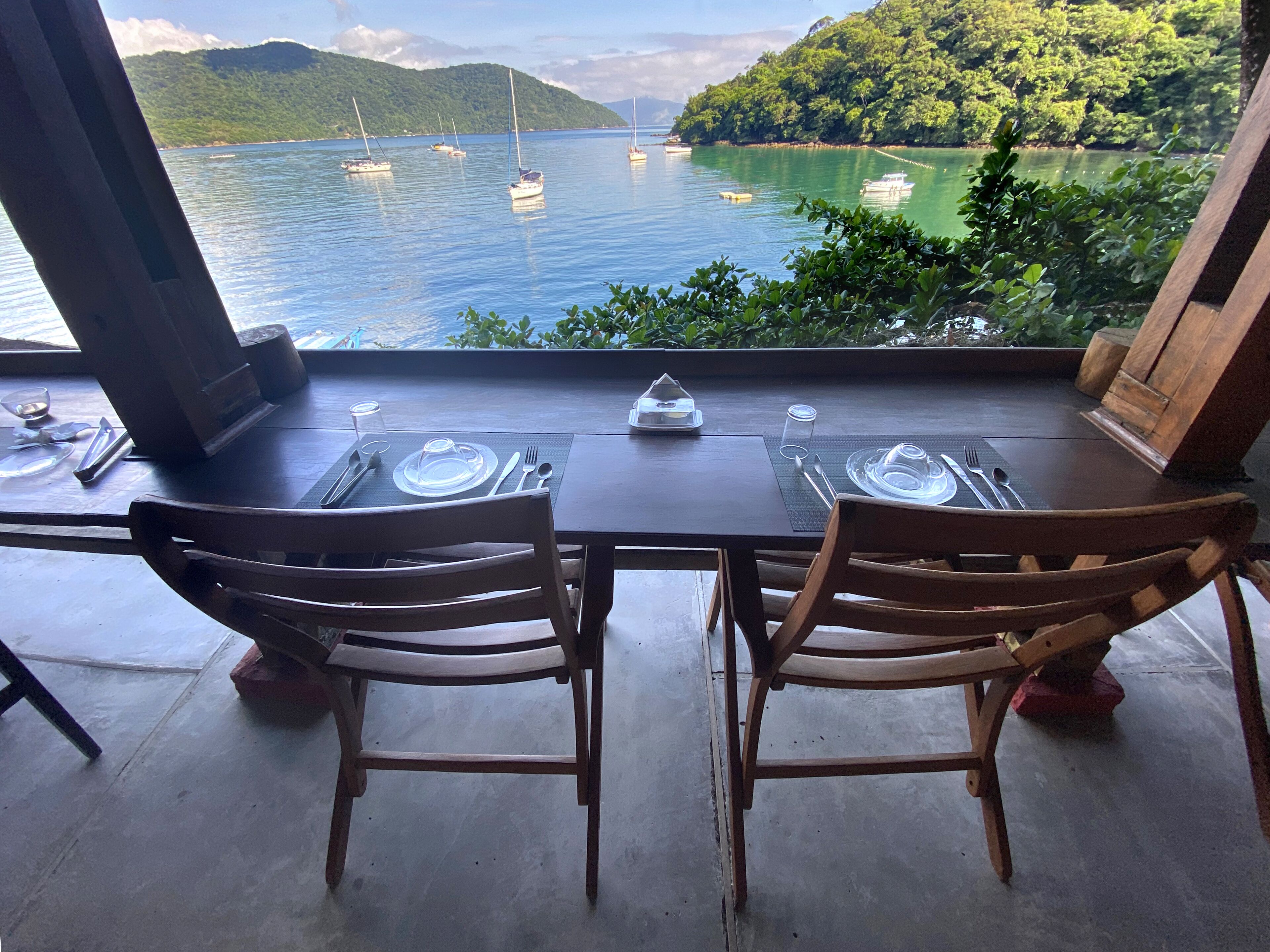 A table set for two with a view of a calm bay with boats and greenery.