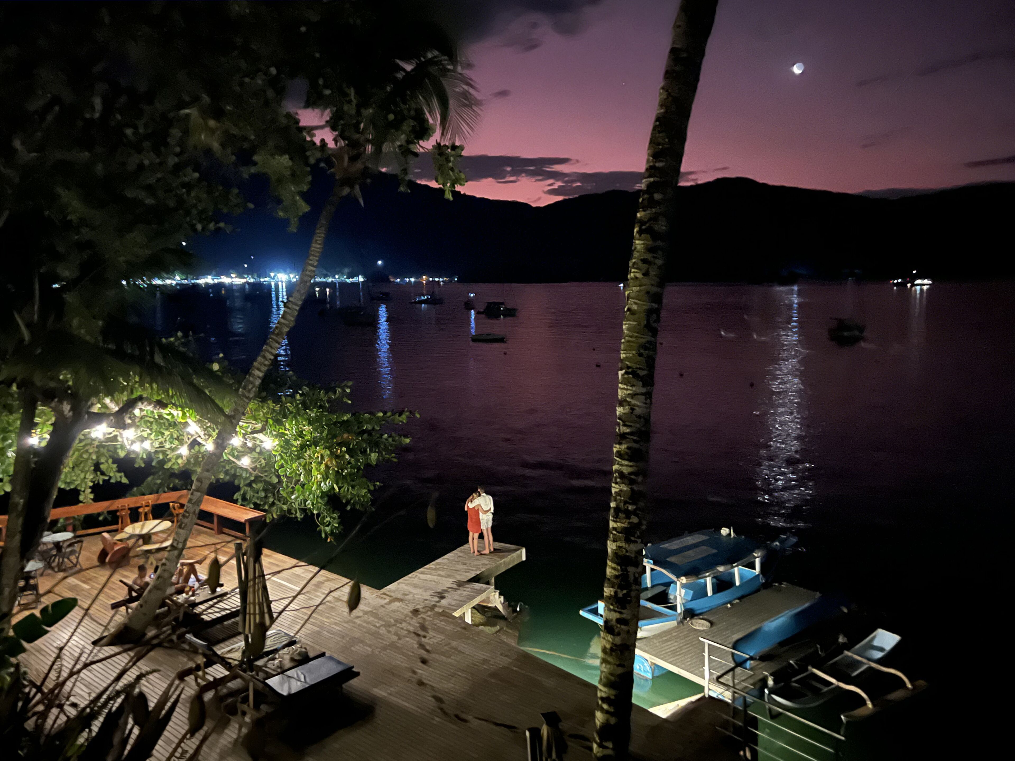 A nighttime view of a dock with boats, a person standing, and a distant cityscape.