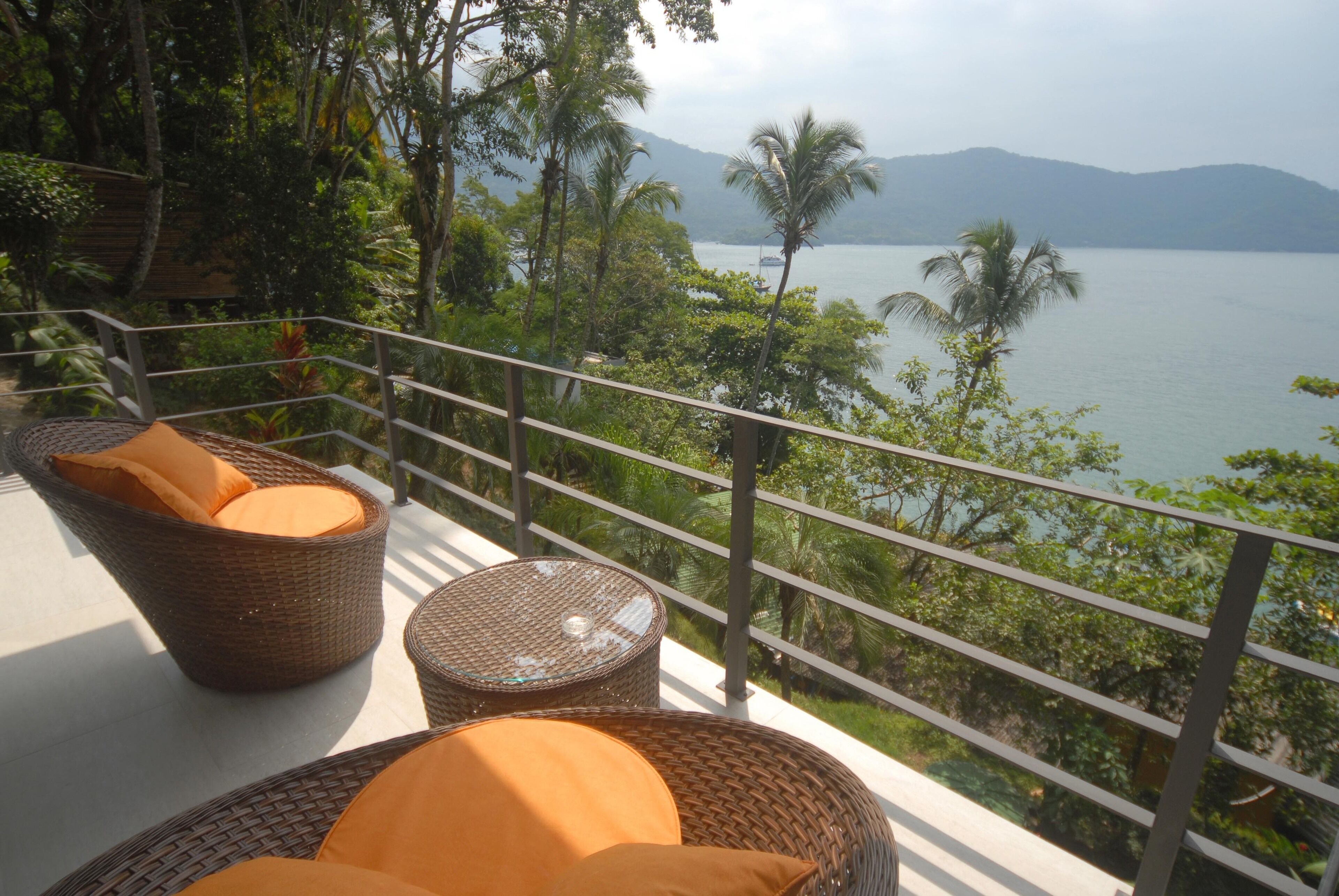 Wicker furniture with orange cushions on a balcony overlooking a lake with palm trees and mountains in the background.