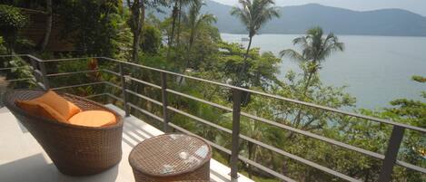 Wicker furniture with orange cushions on a balcony overlooking a lake with palm trees and mountains in the background.