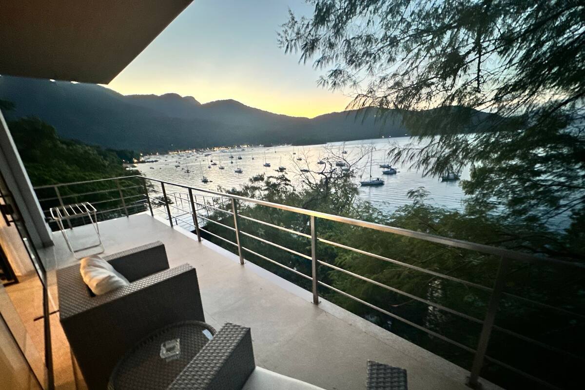 Un balcon avec des sièges d’extérieur, donnant sur un lac où l’on peut voir des bateaux, avec des montagnes en arrière-plan.