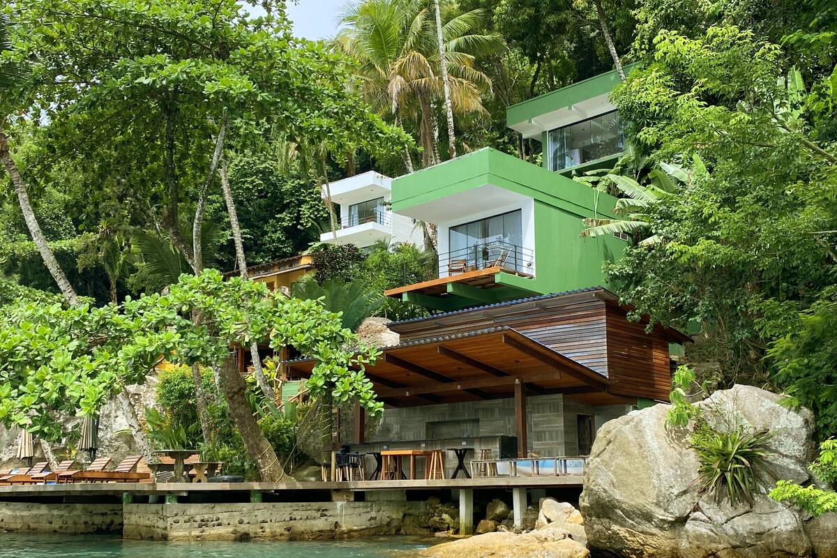 Une maison moderne à plusieurs étages, dotée d’un balcon, entourée d’une végétation luxuriante et bordée d’un plan d’eau.