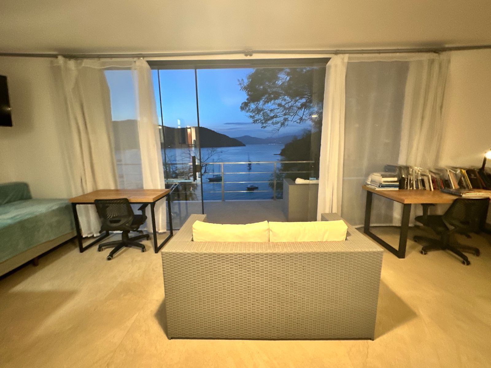 A room with a sofa, a desk, and a chair, with a view of a body of water and hills in the background.