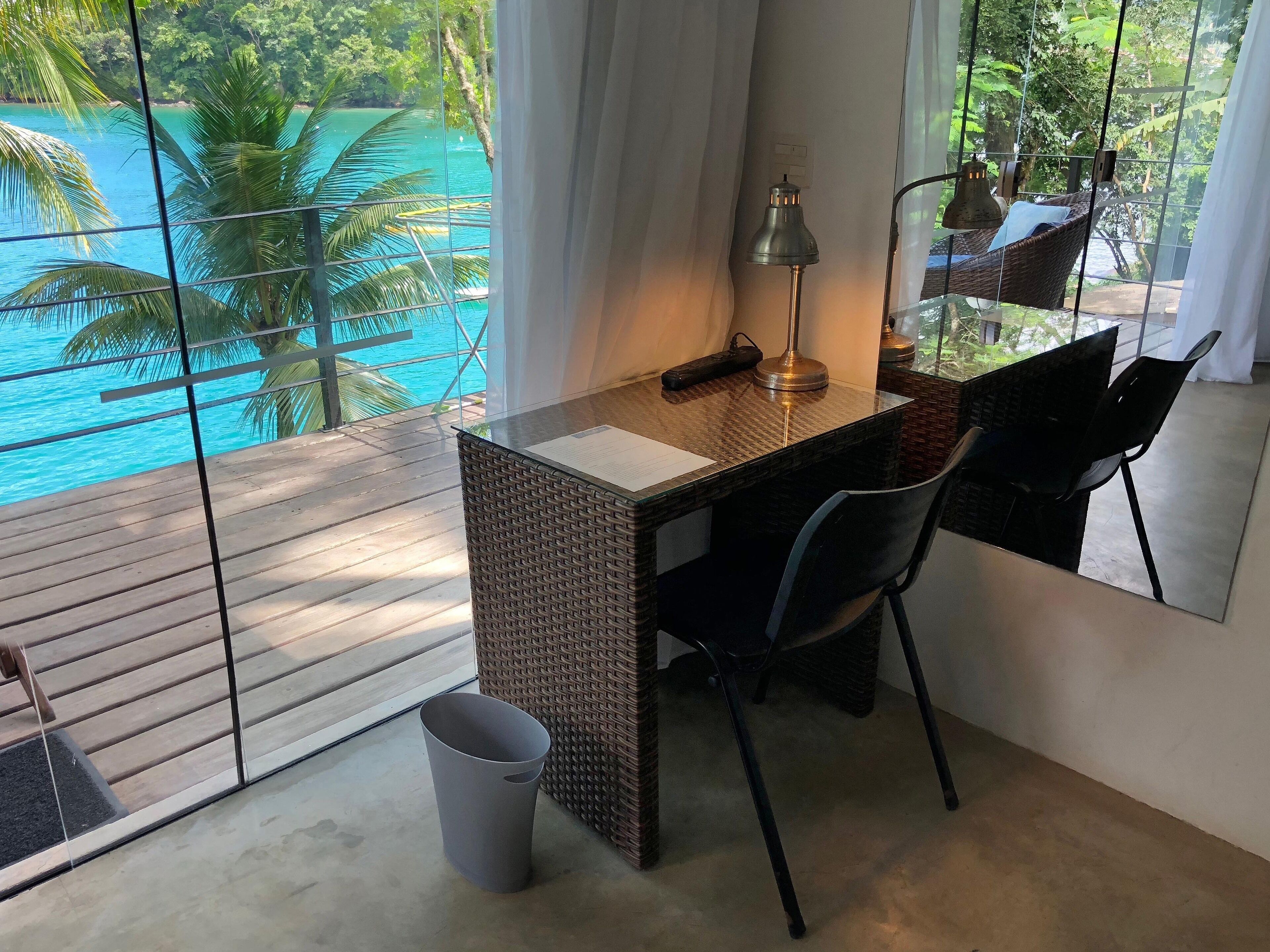 Une pièce avec un bureau en bois, une chaise, une lampe et une vue sur une piscine turquoise et des palmiers.