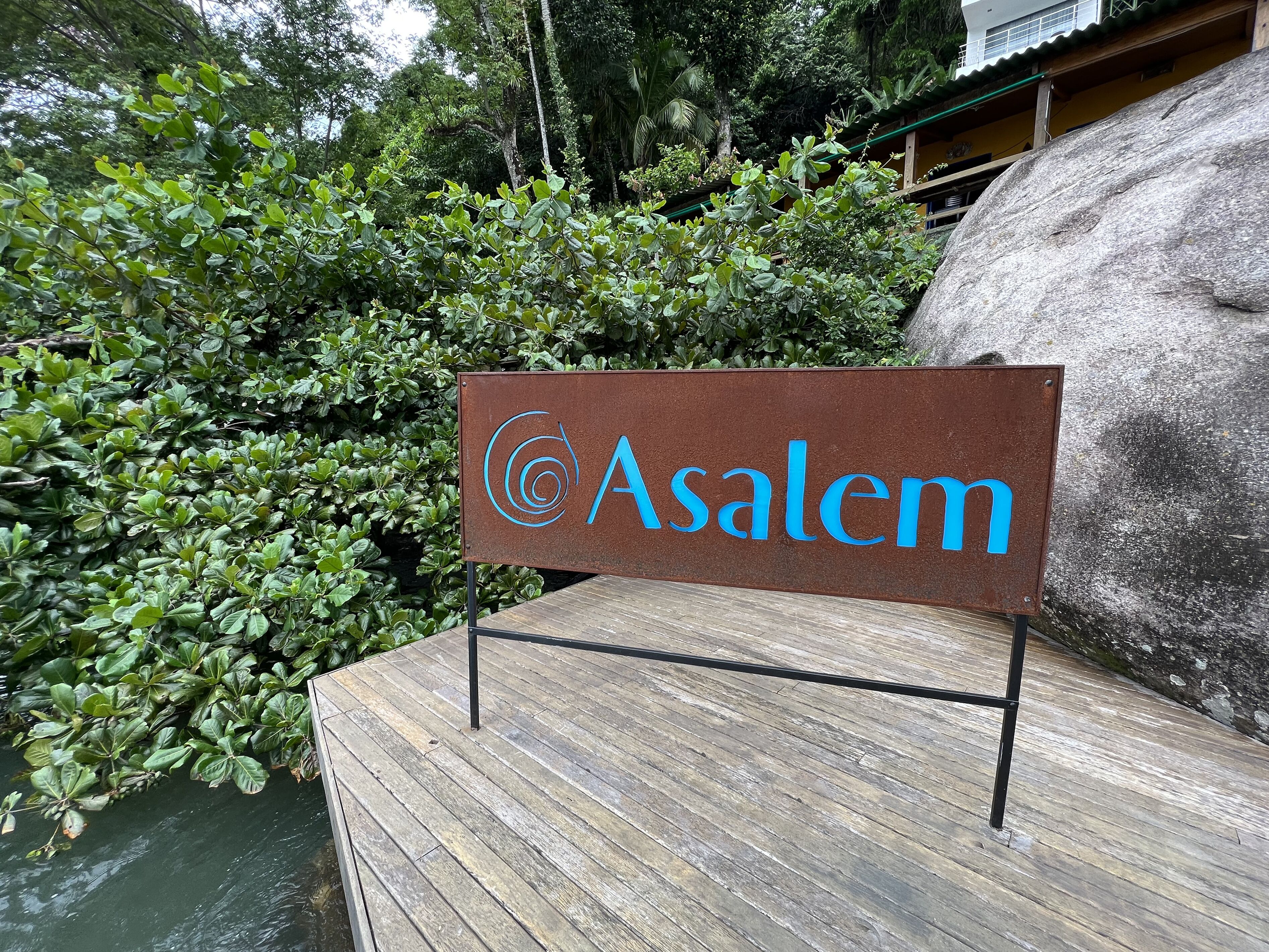 A sign with the name "Asalem" on a wooden platform, surrounded by greenery and a large rock.