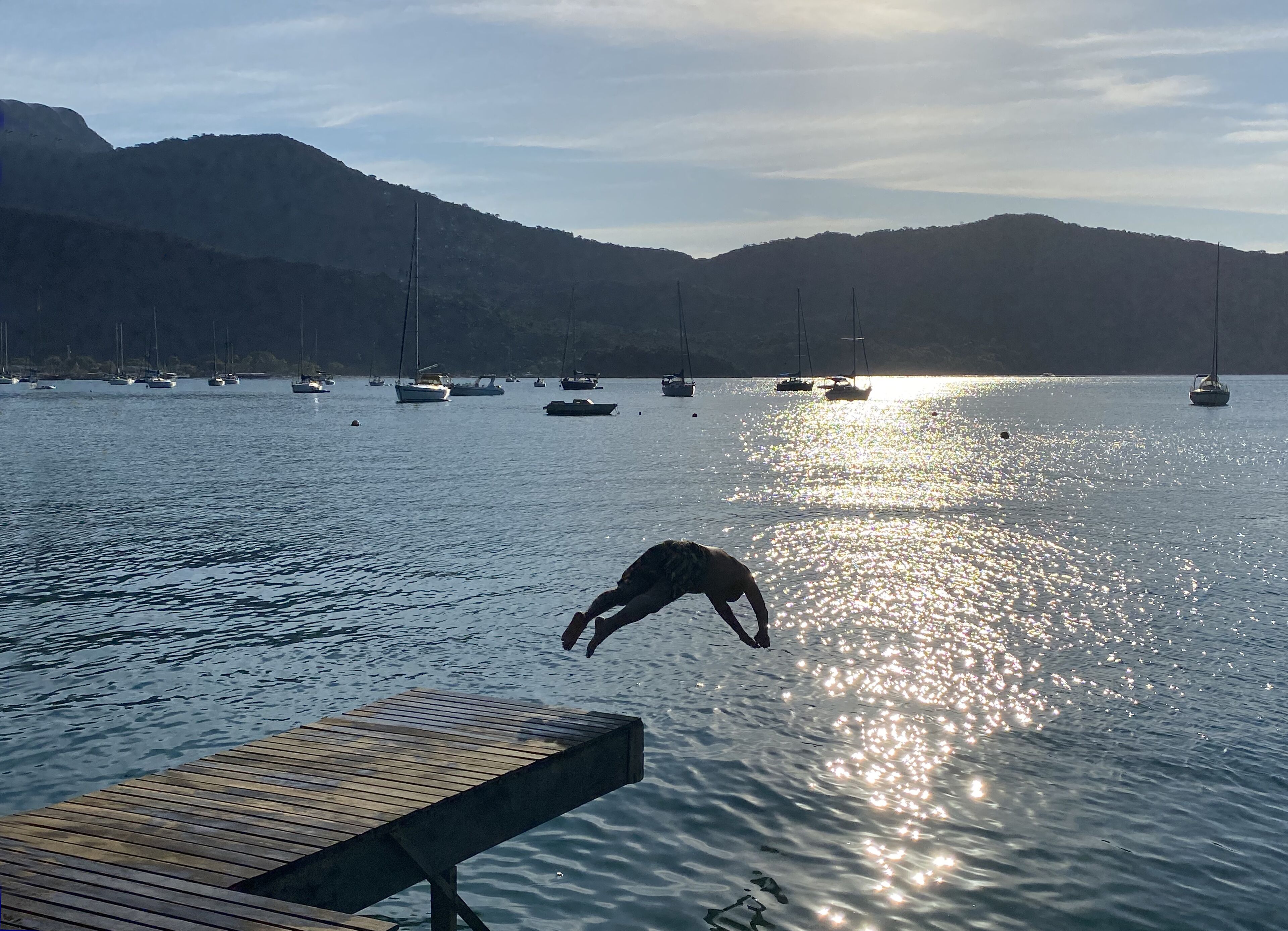 A wooden dock extending into calm water with boats in the background and mountains in the distance.
