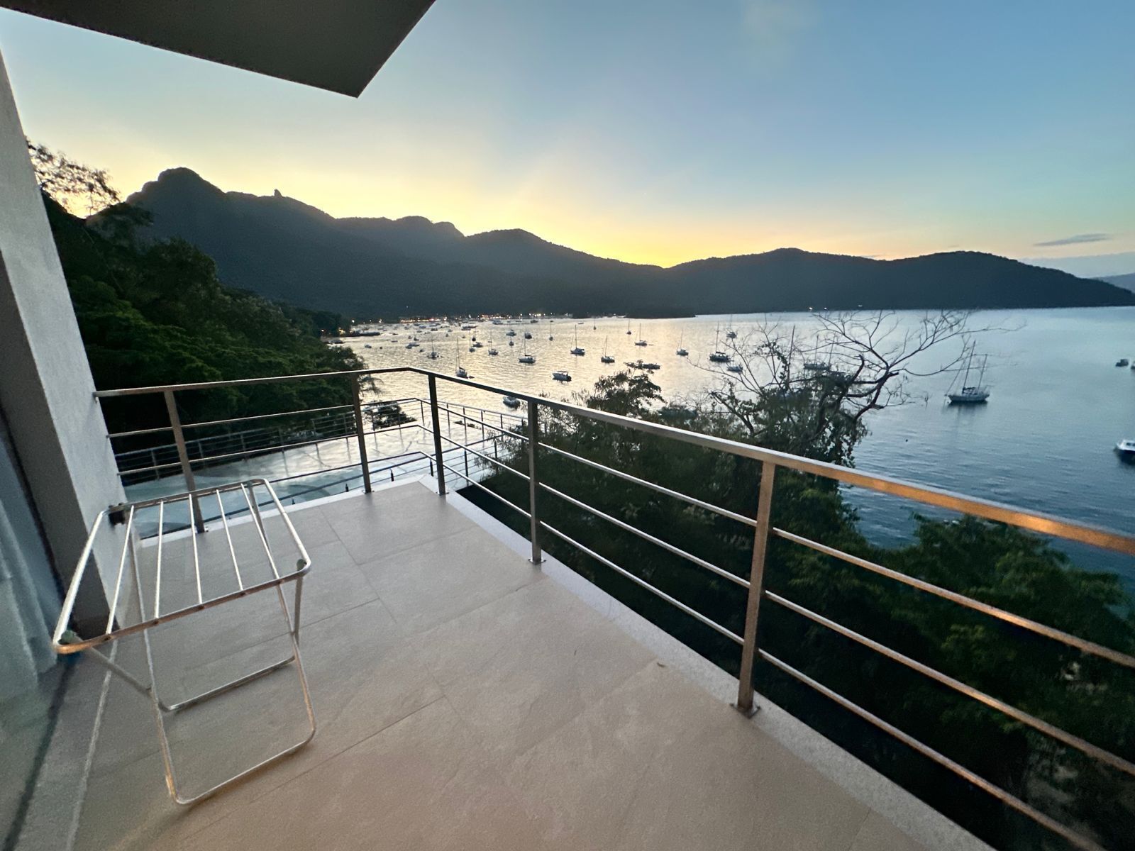 A balcony with a metal railing overlooking a body of water with boats, surrounded by greenery and mountains.