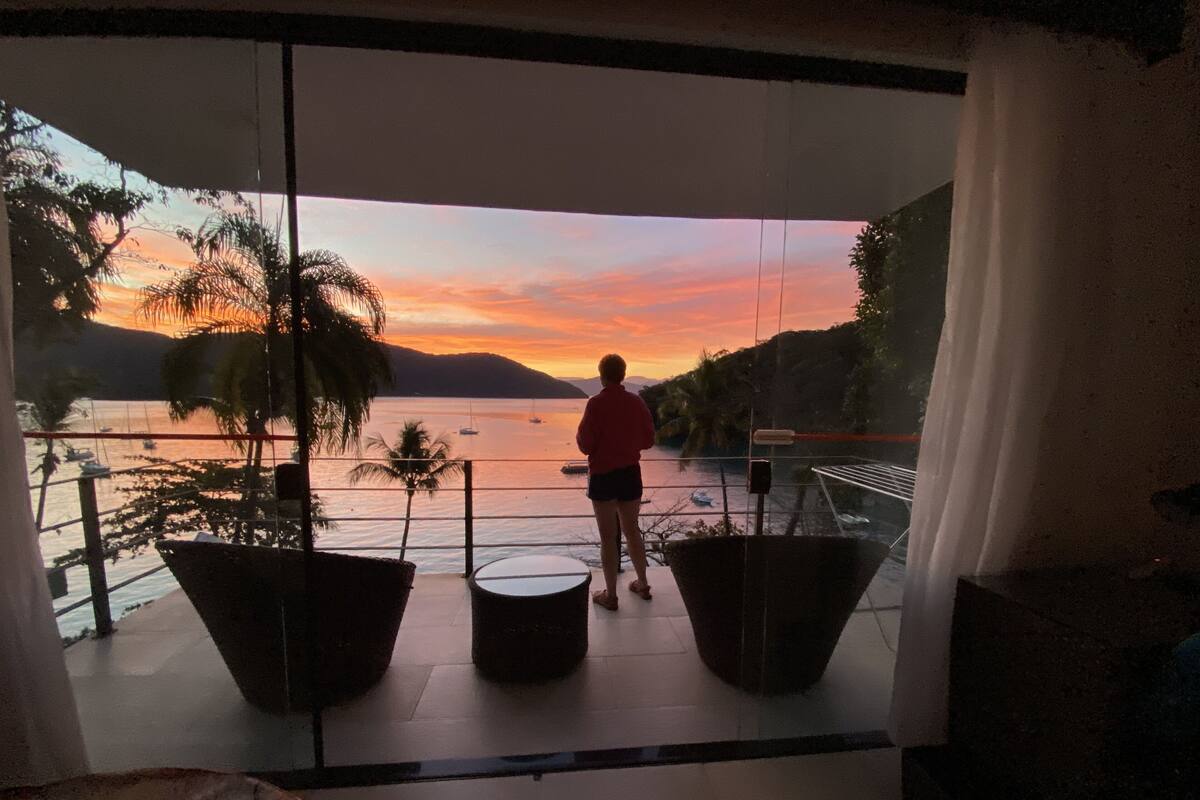 Un balcon avec vue sur un coucher de soleil se reflétant sur l’eau, une personne debout et des plantes en pot.