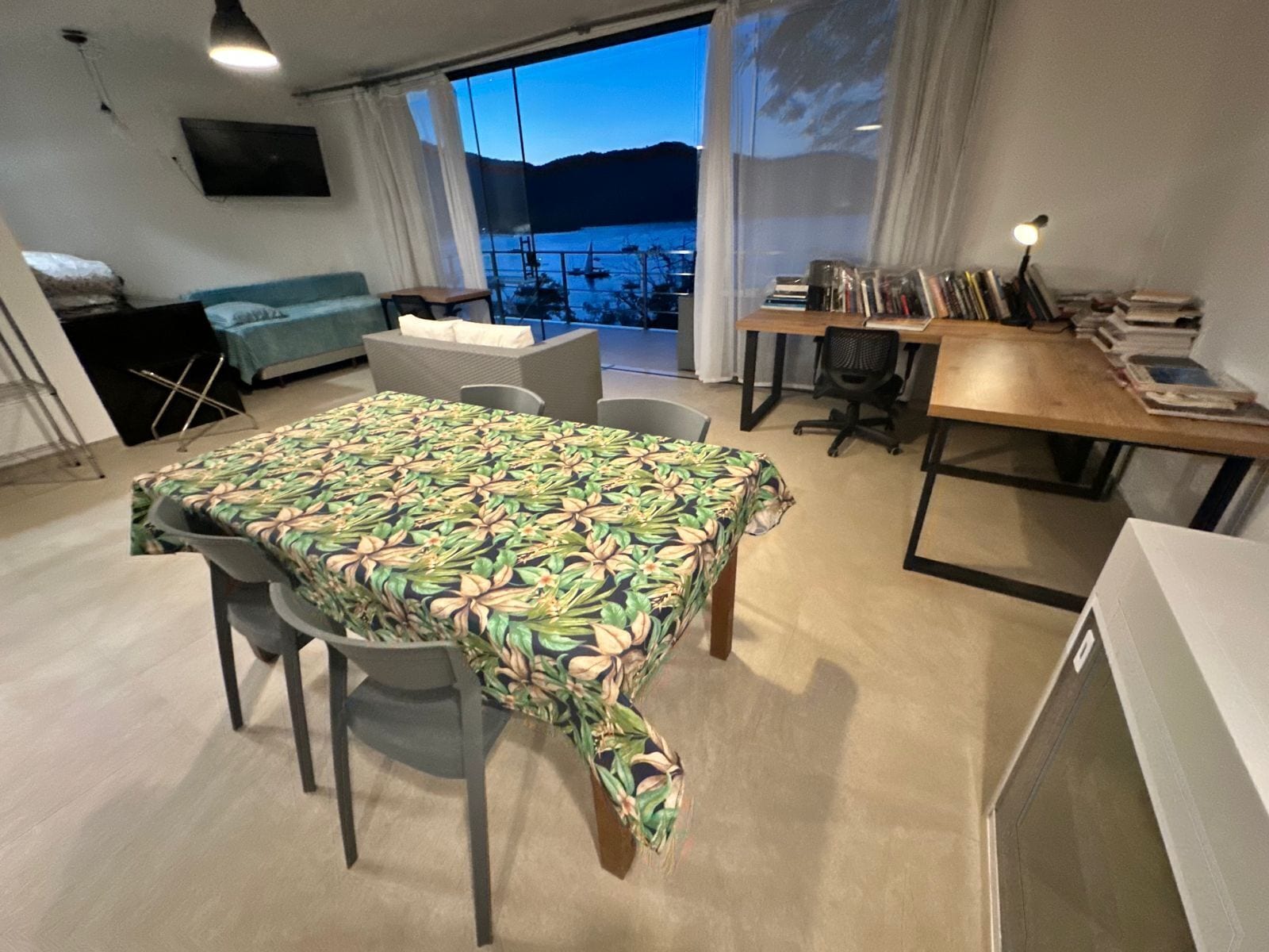 A modern living room with a dining area, a sofa, a TV mounted on the wall, and a view of the sea through large windows.