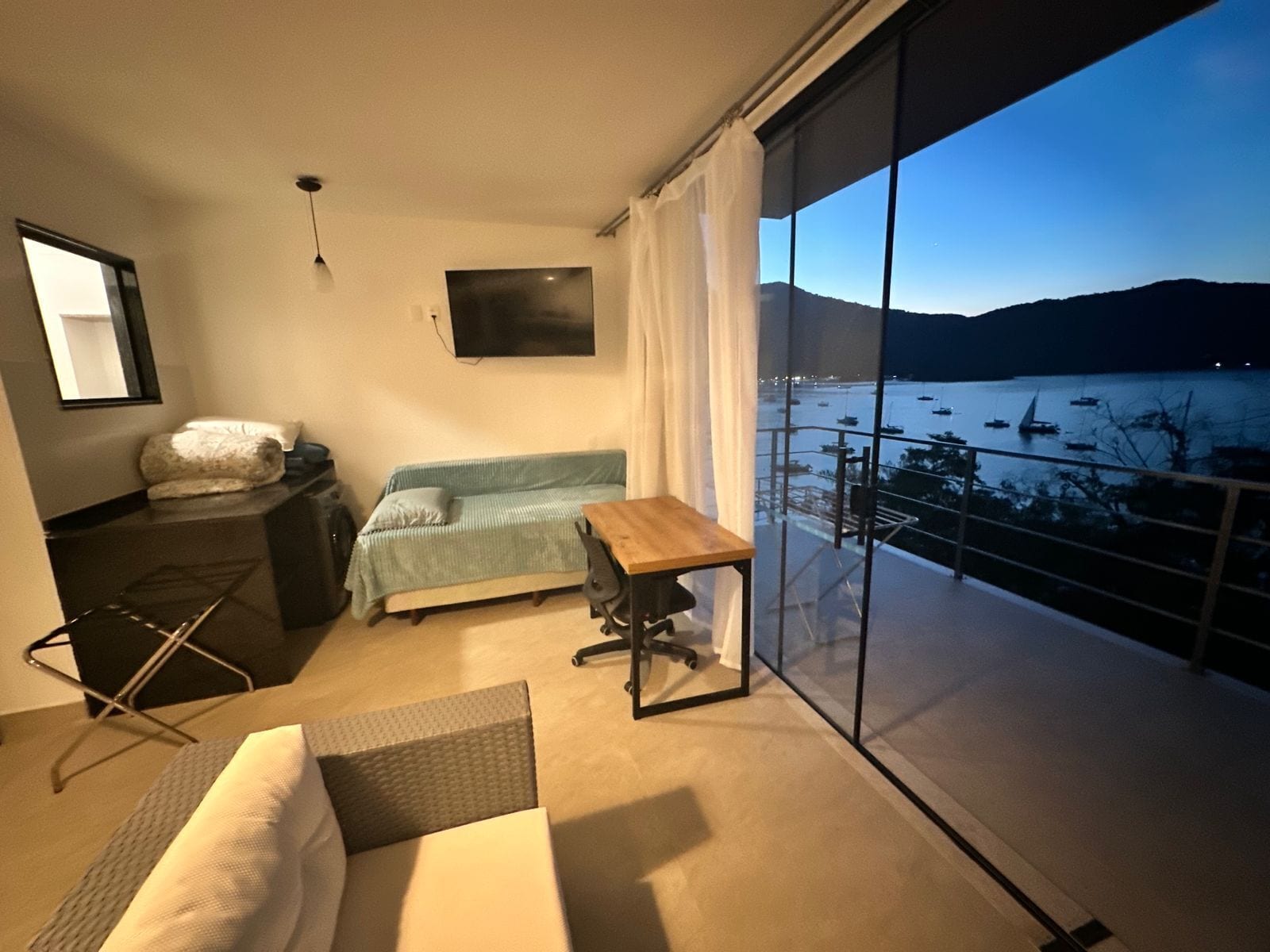 Quarto moderno com cama, escrivaninha pequena, sofá e uma grande janela com vista para um corpo de água e colinas.