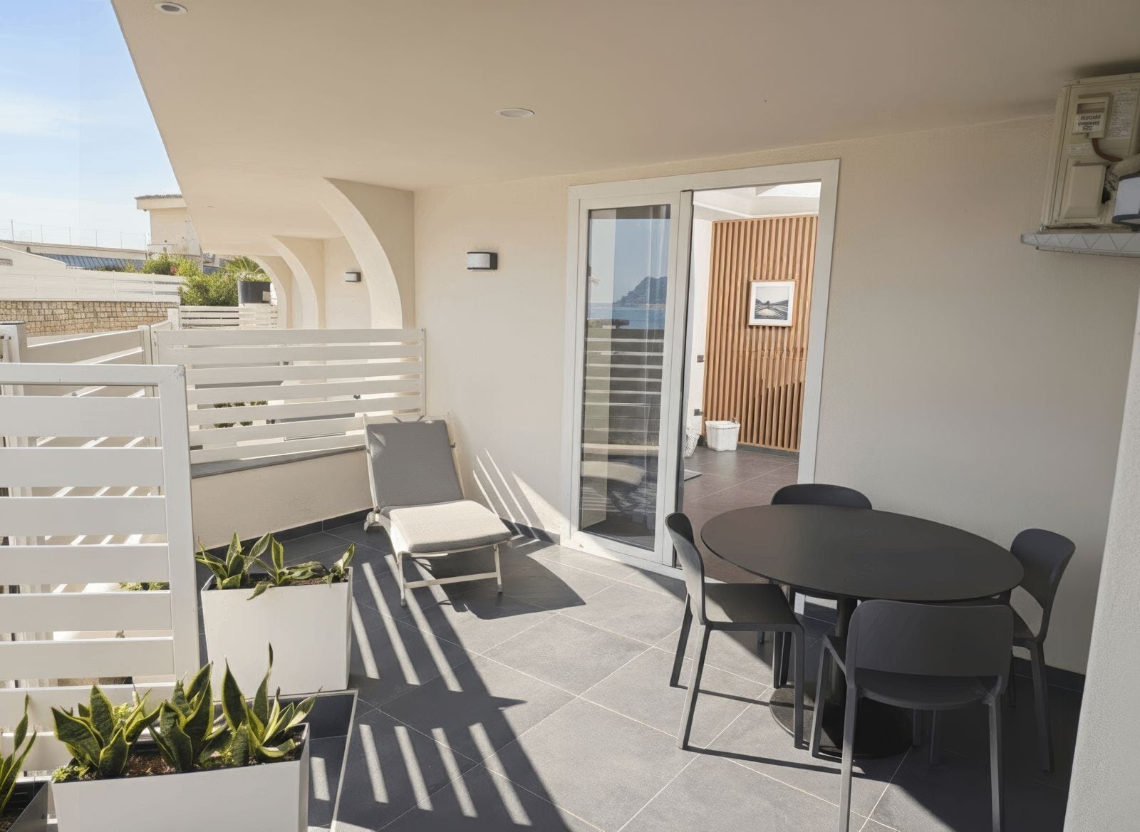 Une terrasse extérieure moderne, dotée d’une porte vitrée donnant sur un balcon, d’une table ronde avec des chaises et de plantes en pot.