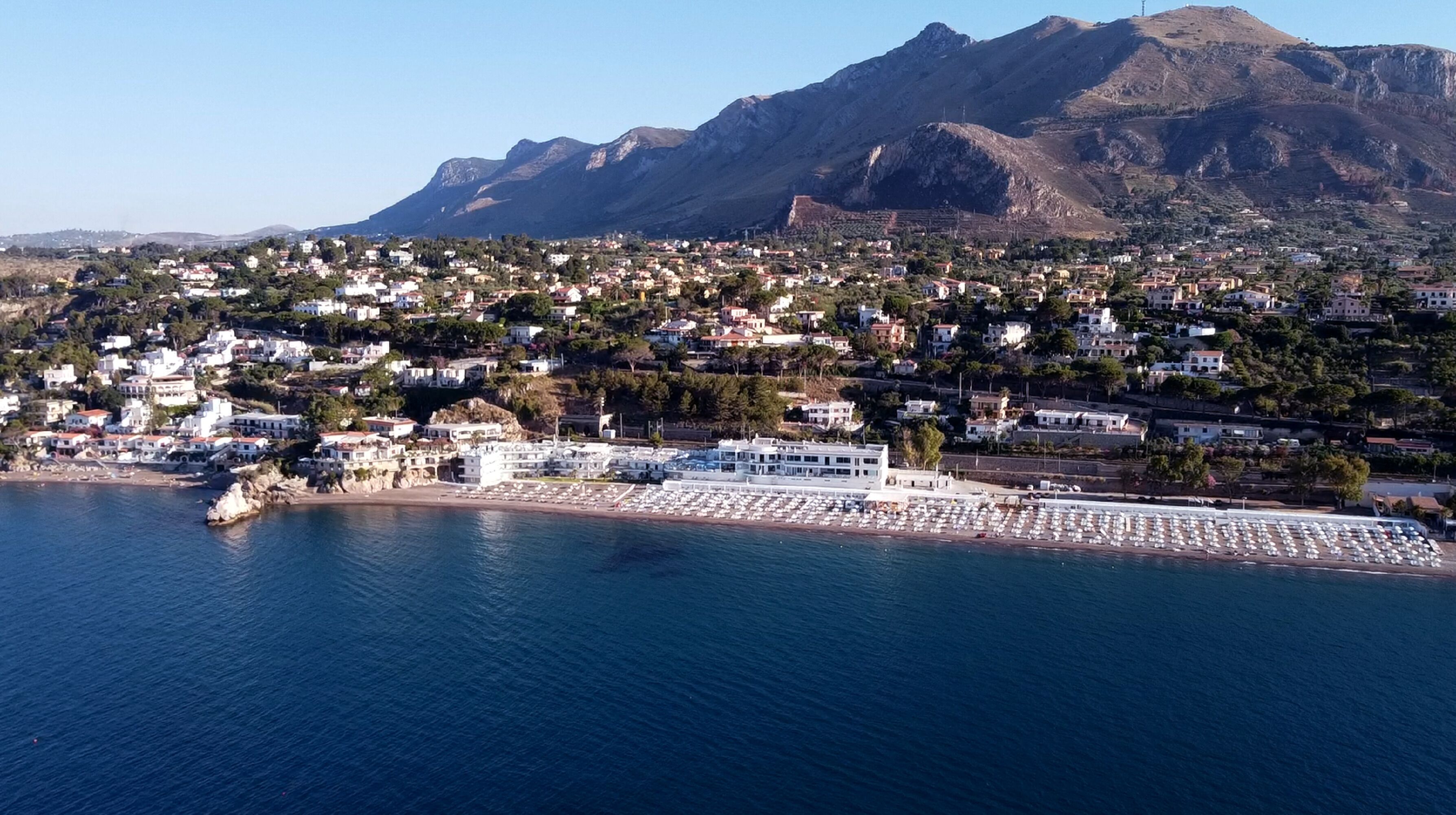 A coastal town with a marina, white buildings, and mountains in the background.