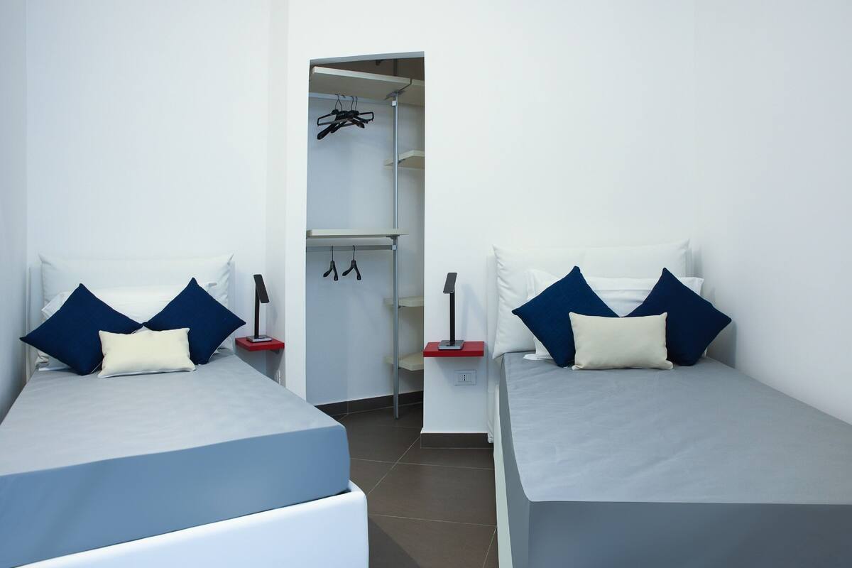 Een kamer met twee bedden, elk voorzien van een kussen en een nachtkastje. Er is een kast met daarin kleding en een plank.