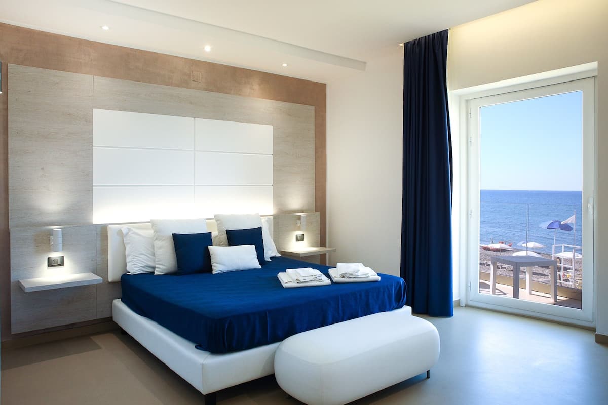 Een moderne hotelkamer met een groot bed, nachtkastjes en uitzicht op de zee door een groot raam.