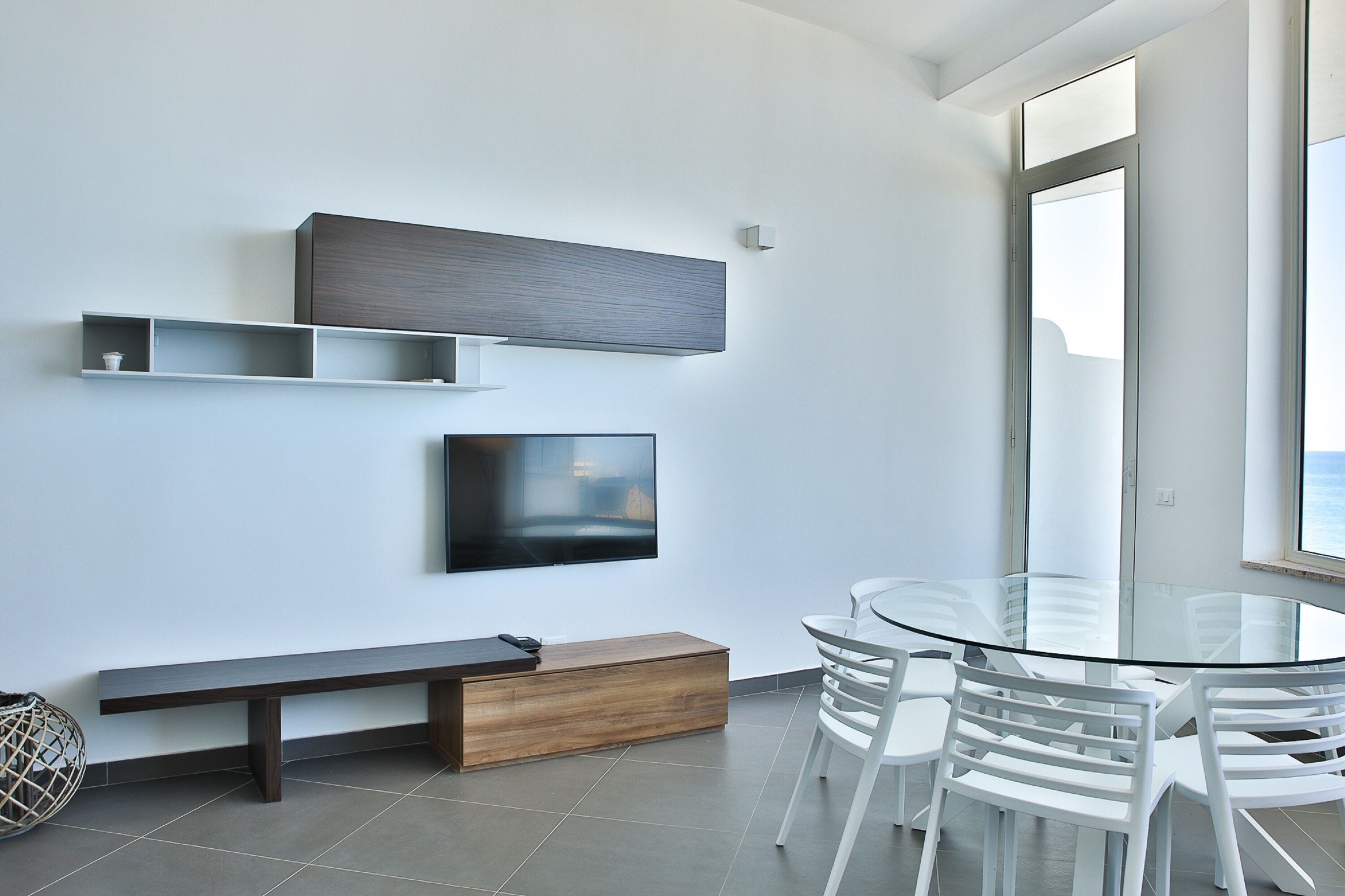 Una habitación moderna con una mesa de vidrio, sillas blancas, un banco de madera, un televisor de pantalla plana y estantes empotrados.