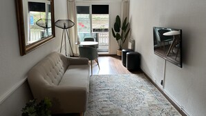 Una sala de estar moderna con un sofá color beige, una mesita redonda pequeña, una lámpara de pie, una planta grande en maceta y un televisor montado en la pared.