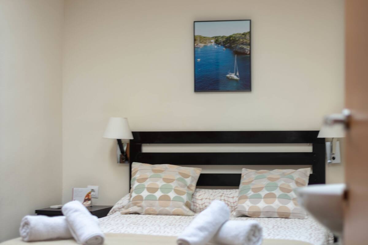 A neatly made bed with rolled towels, a nightstand with a lamp, and a painting of a coastal scene above the bed.