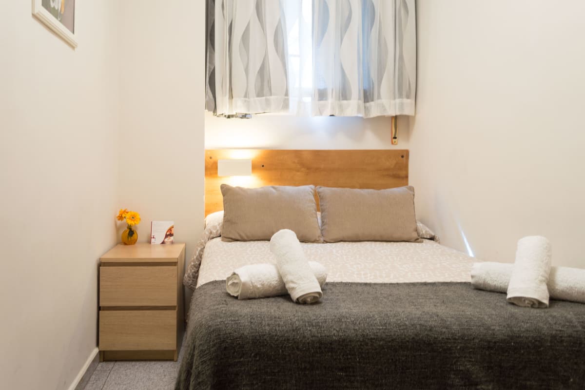 A neatly made bed with a dark blanket, two pillows, and rolled towels on a wooden nightstand beside it.