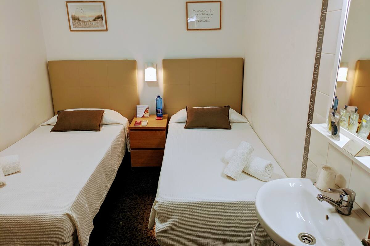 A hotel room with two beds, a nightstand with a water bottle, a mirror, and a sink.