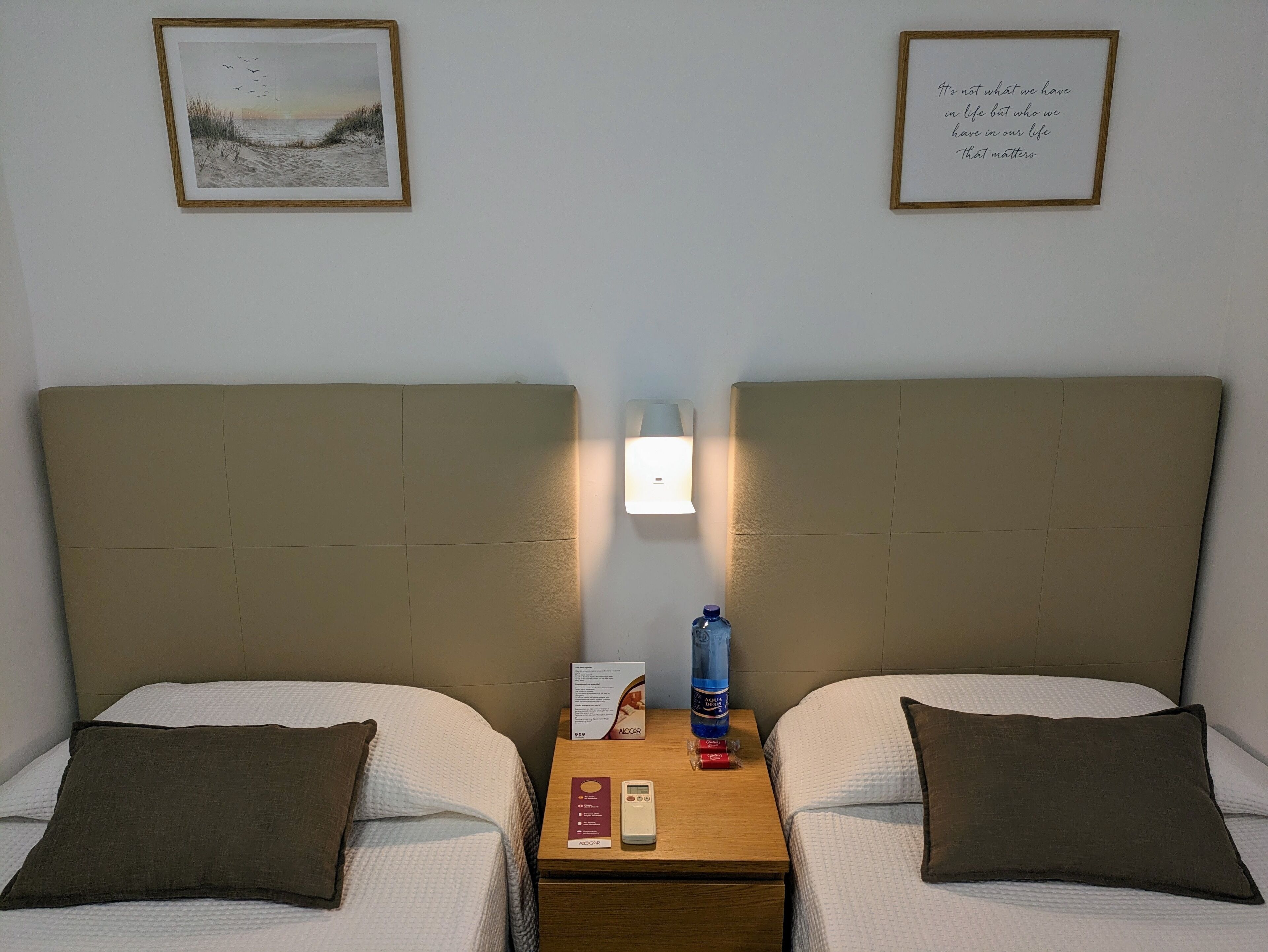Habitación de hotel con dos camas, una mesita de noche con lámpara, una botella de agua y una cita enmarcada en la pared.