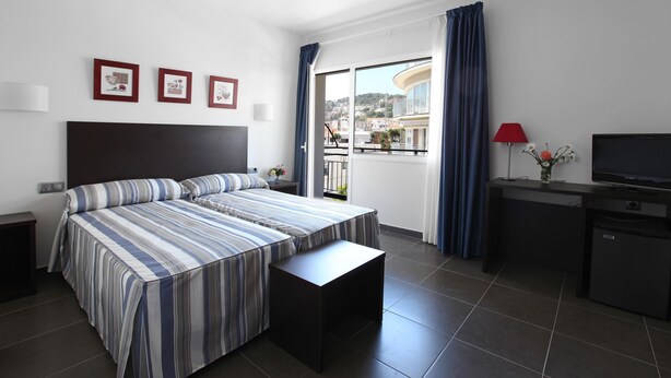 A hotel room with a bed, a desk, a TV, and a balcony with a view of buildings. - Hotel Marblau Tossa (Tossa de Mar)