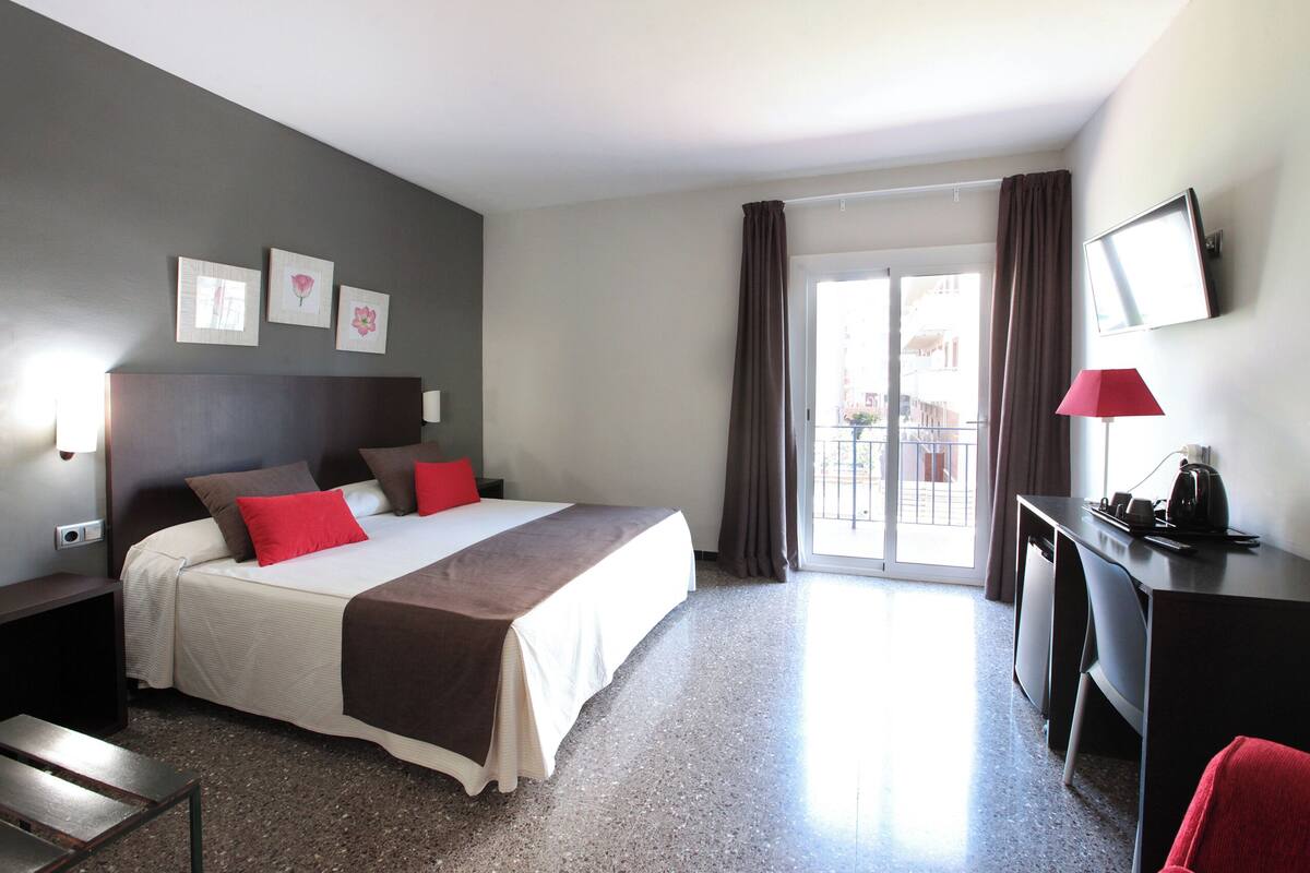 Habitación de hotel con cama, escritorio, silla, televisión y balcón con cortinas.