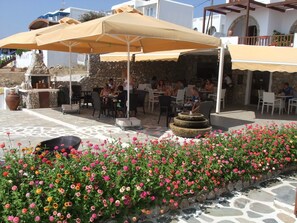 An outdoor seating area with stone walls, tables, and chairs under large umbrellas. - Castillio Hotel (Astypalaia)