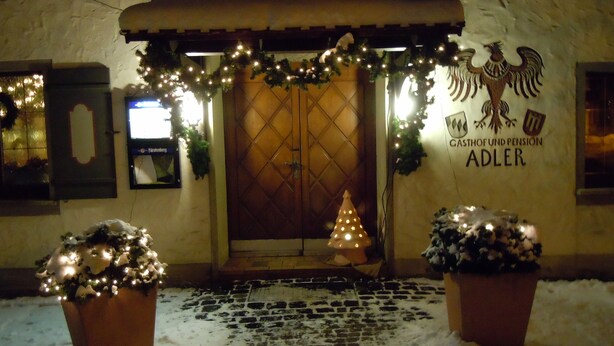L’entrée d’un immeuble avec une porte en bois, un sol recouvert de neige et des décorations de Noël, notamment un sapin illuminé et des guirlandes.
