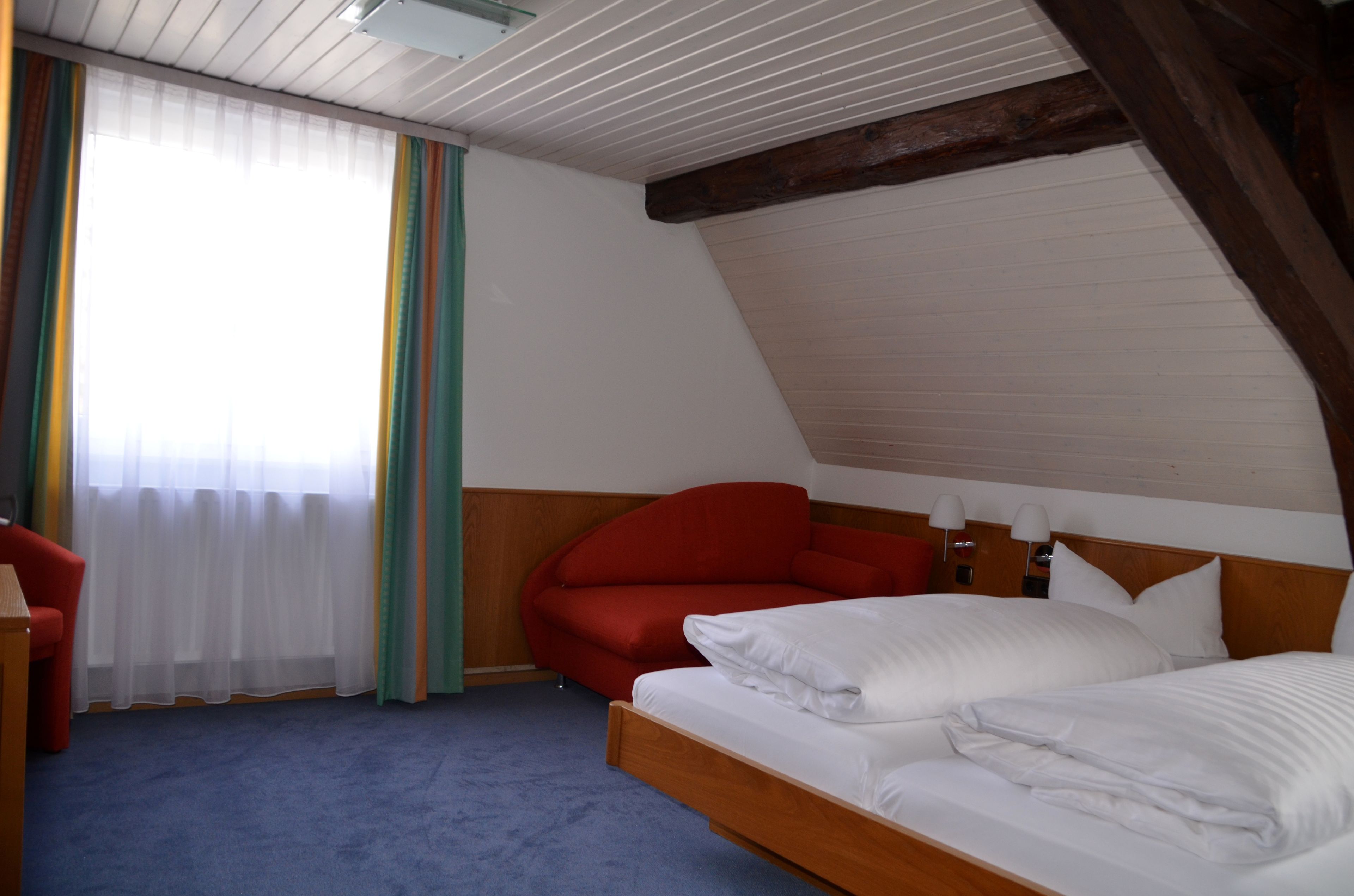 Ein Hotelzimmer mit Holzboden, einem Bett mit weißen Leinen, einem roten Sessel und einem Schreibtisch mit Lampe.