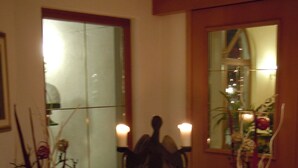 Un couloir avec un grand miroir reflétant une pièce, une statue entourée de bougies et deux grands arrangements floraux.