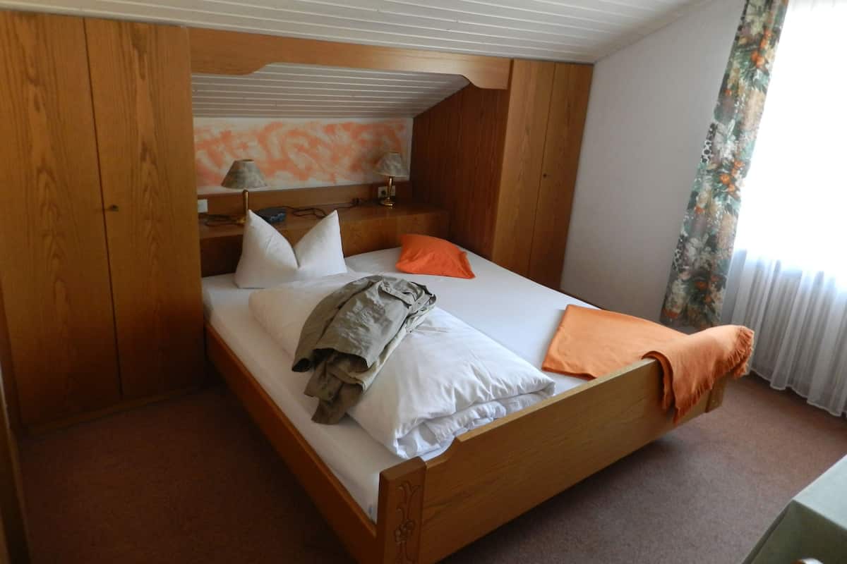 Een eenpersoonsbed met een houten frame, witte beddengoed en een deken. Links een houten garderobe en rechts een raam met gordijnen.