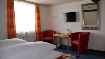Ein Hotelzimmer mit zwei Betten, einem Fernseher, einem kleinen Tisch mit zwei Stühlen und einem Fenster mit Vorhängen.