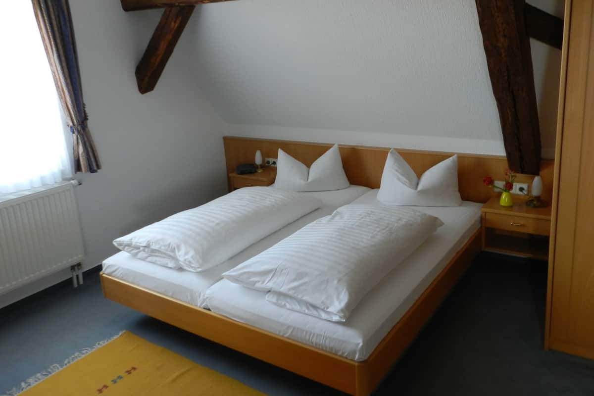Een tweepersoonsbed met witte lakens, een houten hoofdbord, een nachtkastje met een lamp en een raam met gordijnen.