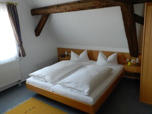 Una cama doble con ropa blanca, cabecera de madera, mesita de noche con lámpara y ventana con cortinas.