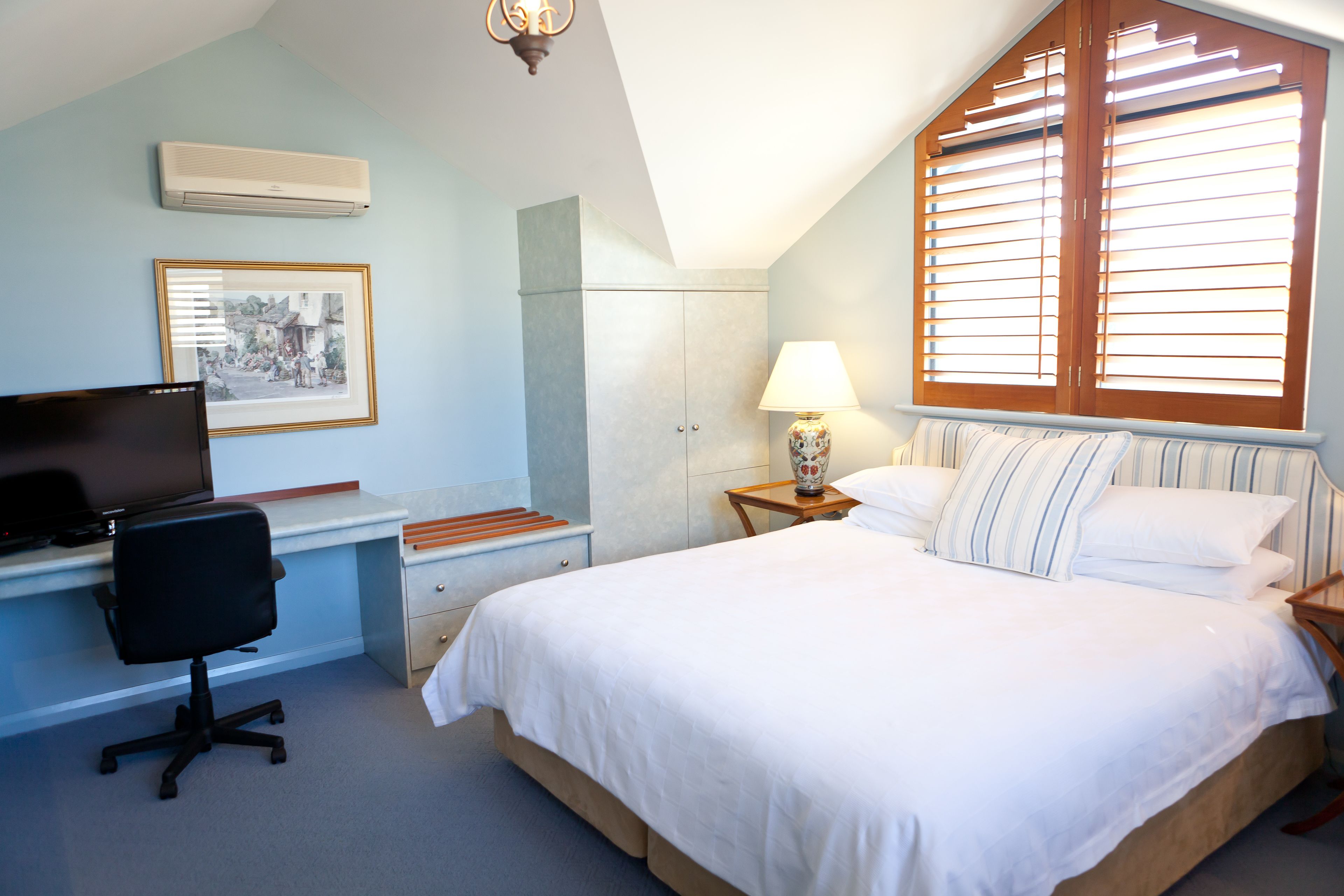 A bedroom with a bed, a desk with a chair, a TV, a lamp, and a window with blinds.