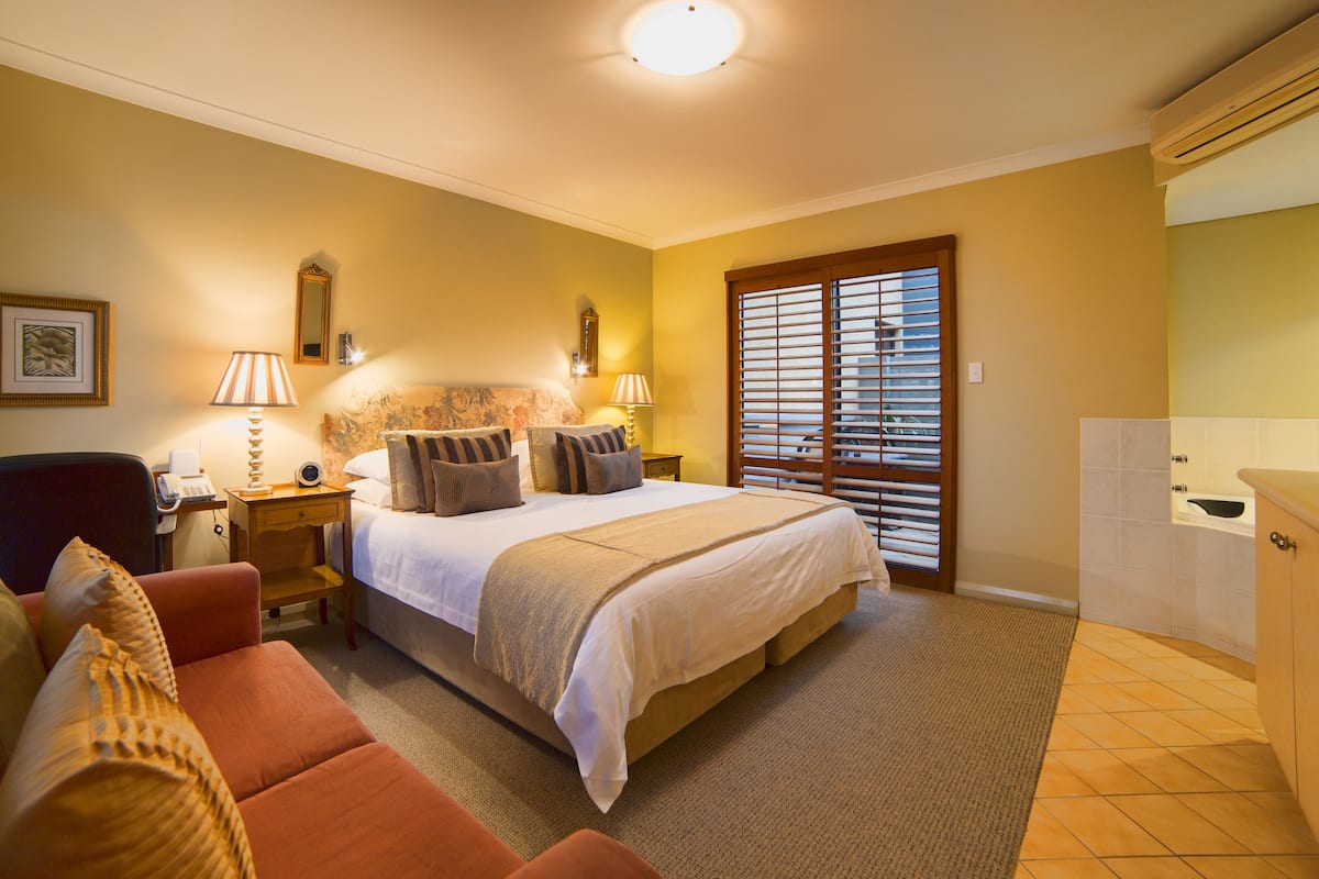 A hotel room with a bed, a sitting area with a chair and a table, a bathroom with a bathtub, and a window with blinds.