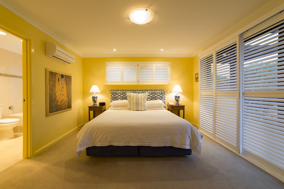 A bedroom with a bed, two bedside lamps, a window with blinds, an air conditioning unit, and a painting on the wall.