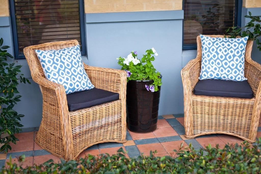 Two wicker chairs with patterned cushions, a potted plant with white flowers, and a tiled floor.