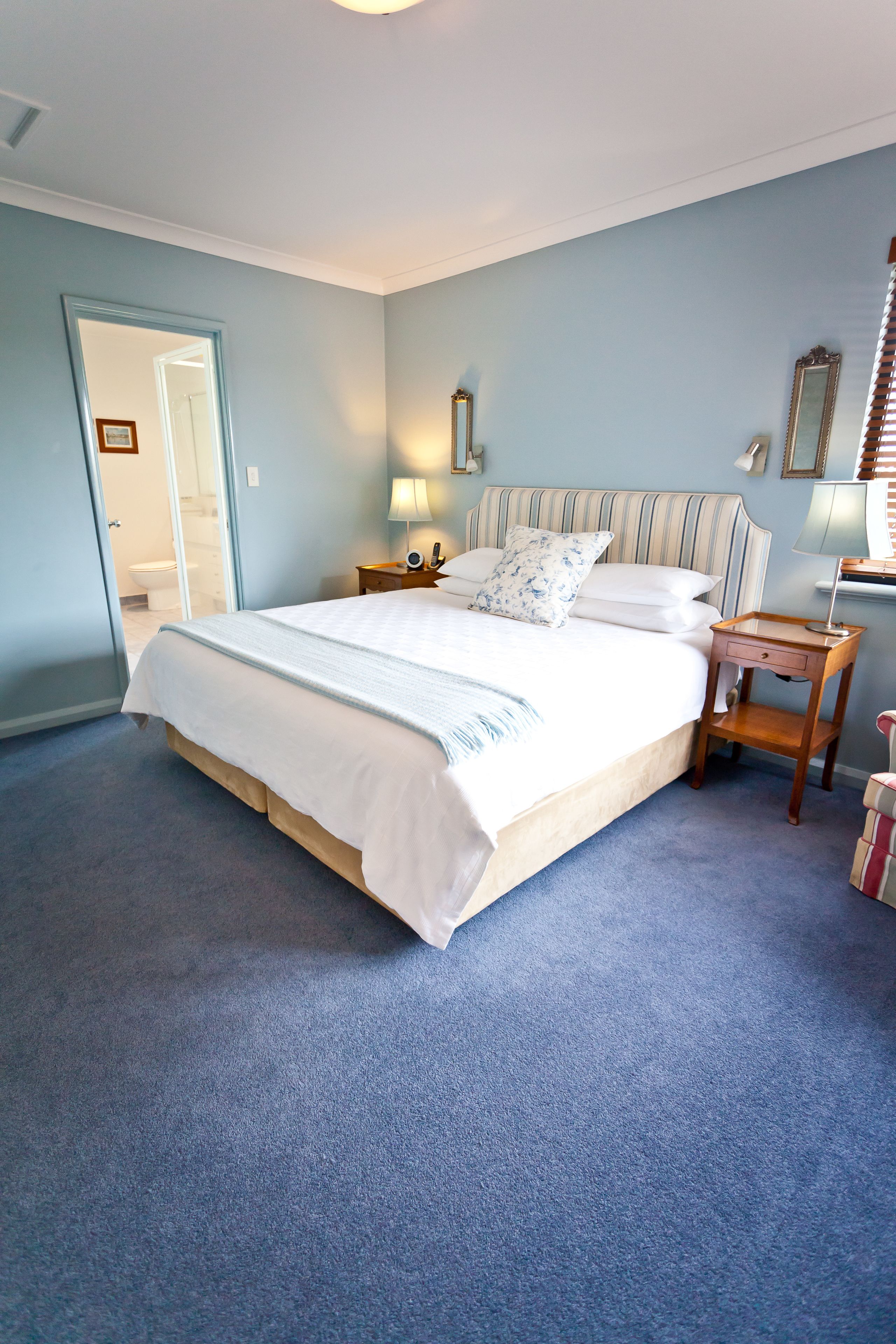 A neatly made bed with white linens and a striped headboard, a bedside table with a lamp, and a mirror on the wall.