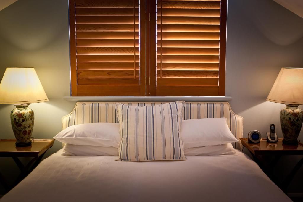 Une chambre d’hôtel avec un lit, une tête de lit à rayures, deux lampes de chevet et une fenêtre avec des stores.