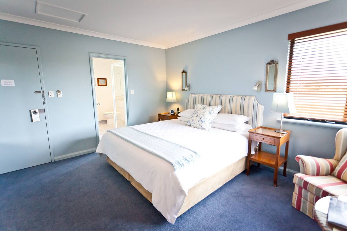 A bedroom with a bed, a nightstand, a chair, a window with blinds, and a door.