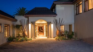 A well-lit entrance with a tiled pathway leading to a covered entryway, flanked by large vases and plants. - Manor 38 (Gqeberha (Port Elizabeth))