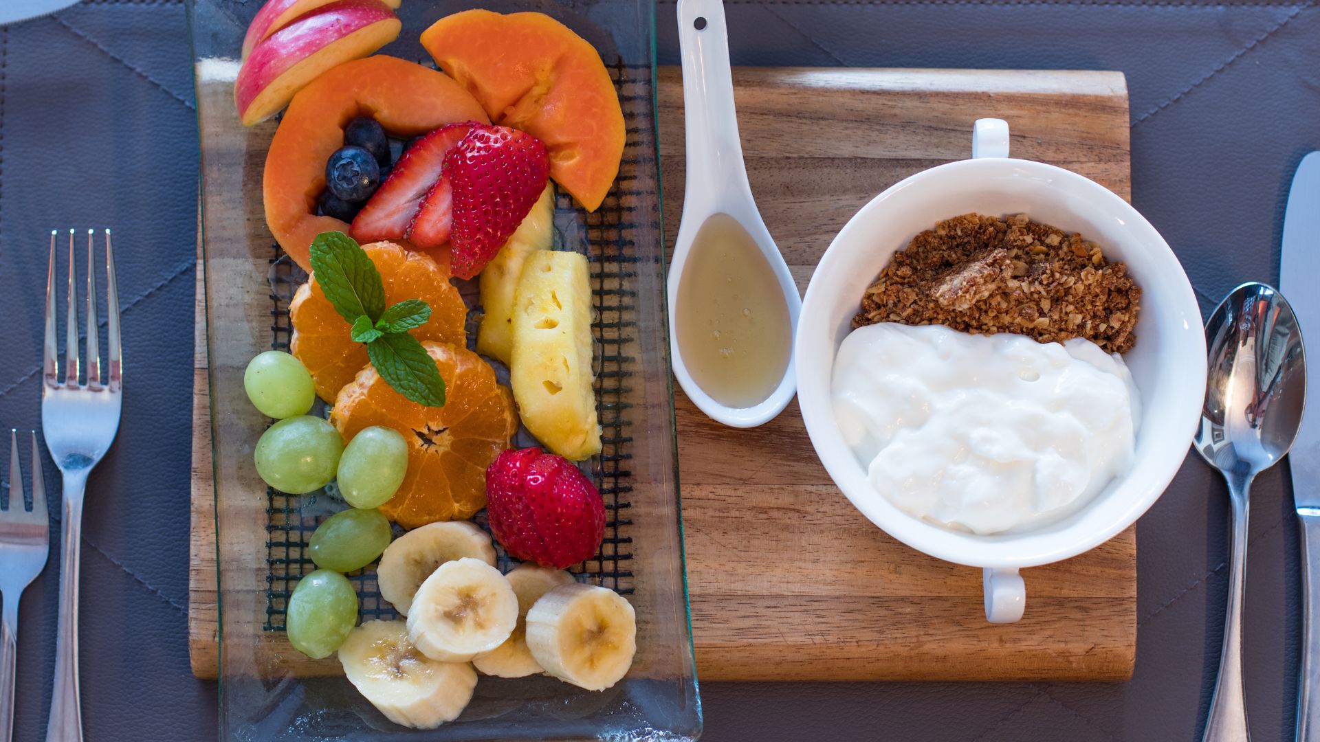 Een ontbijt met een fruitschaal, yoghurt, granola en een honinglepeltje op een houten plank.