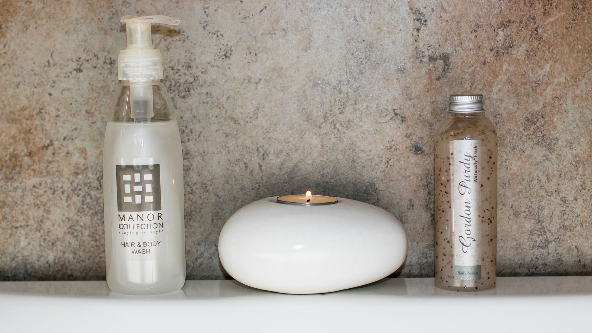Un estante de baño con una botella de jabón de pelo y cuerpo, una vela y una botella de exfoliante corporal.